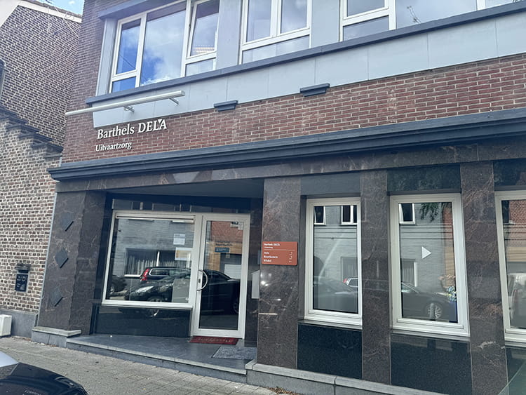 Aanzicht buitengevel met witte glazen deur en vier witte ramen waarboven Uitvaartzorg Barthels DELA staat